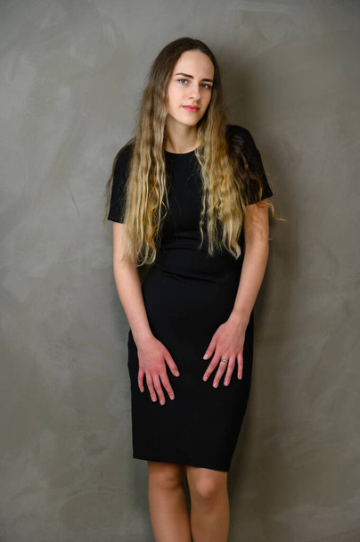 Vertical studio photo on gray background of a cute caucasian teenager girl in a black dress with long hair - Valokuva, kuva