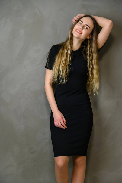 Vertical photo of a cute caucasian teenager girl in a black dress with long hair in the studio on a gray background - 写真・画像