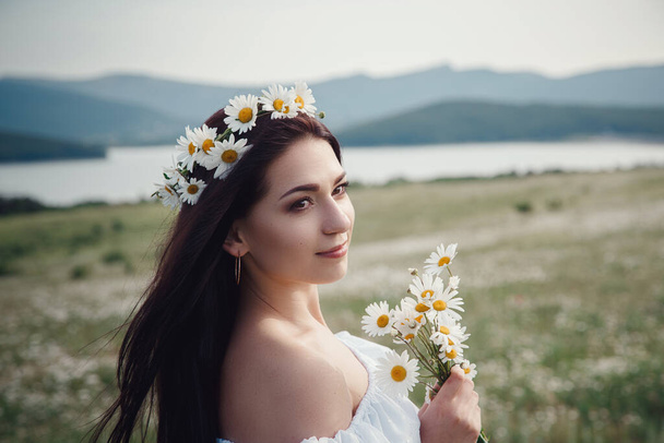 Hermosa mujer morena joven en un vestido blanco y corona de flores disfrutando de la primavera en un campo de margaritas. Idea y concepto de salud, hermoso cabello sano y piel, falta de alergias
 - Foto, imagen