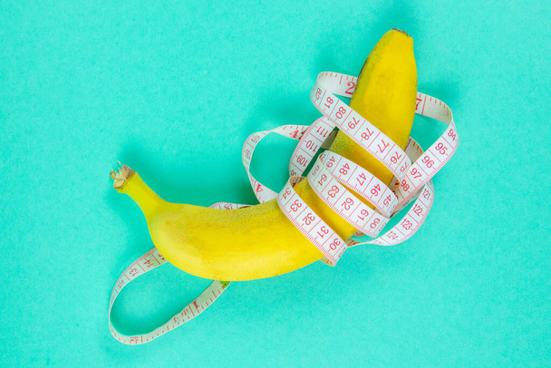 Μπανάνα με ταινία για τη μέτρηση της μορφής. Ο χάρακας του εκατοστού περιστρέφεται γύρω από τα φρούτα. Ταινία τυλιγμένη γύρω από μπανάνα απομονωμένη σε φόντο μέντας. Απώλεια βάρους, υγιεινή διατροφή και λεπτή έννοια του σώματος. - Φωτογραφία, εικόνα