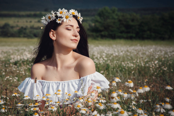 Beautiful young brunette woman in a white dress is enjoying spring in a field of daisies. Summer countryside concept. Close to nature vacation. Woman and daisies. Summer light - Photo, image