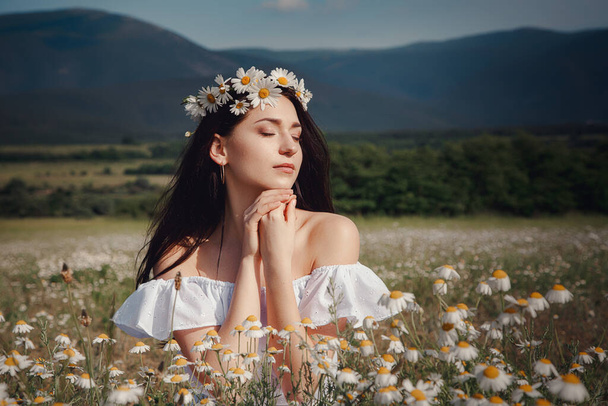 Beautiful young brunette woman in a white dress is enjoying spring in a field of daisies. Summer countryside concept. Close to nature vacation. Woman and daisies. Summer light - Photo, Image