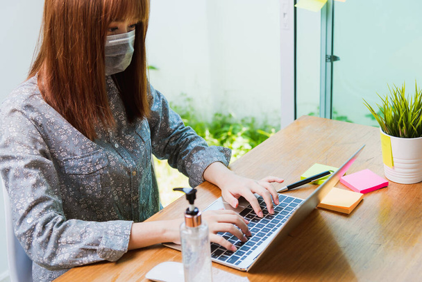 Asian Business beautiful young woman work from home office he wearing a protective mask with disinfectant gel beside her working laptop computer online quarantines disease coronavirus or COVID-19 - Fotografie, Obrázek