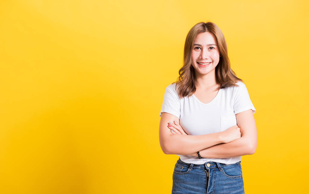 Asian Thai happy portrait beautiful cute young woman standing wear t-shirt her smile confidence with crossed arms looking to camera isolated, studio shot on a yellow background and copy space - 写真・画像