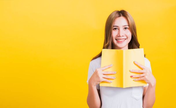 Portrait Asian Thai beautiful happy young lifestyle woman stands holding yellow book or diary her looking to camera, studio shot isolated on yellow background, with copy space - Foto, immagini
