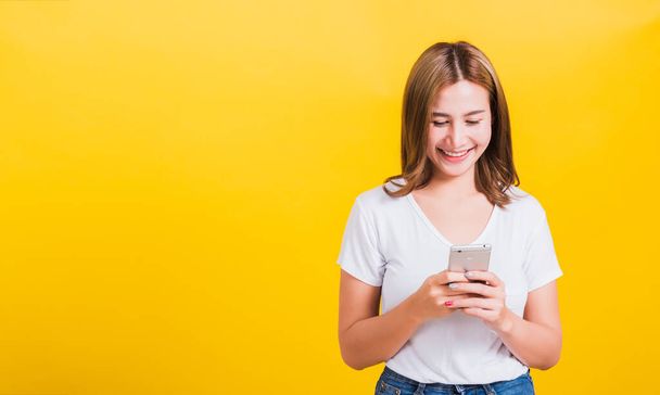Asian Thai portrait happy beautiful cute young woman smile standing playing game or writing SMS message on smartphone looking to the phone, studio shot isolated on yellow background with copy space - Zdjęcie, obraz