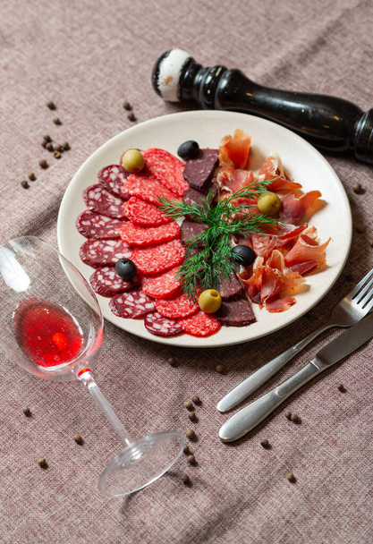 sausage sliced with basturma, olives and herbs in a large ceramic plate on a coffee color tablecloth - Photo, Image