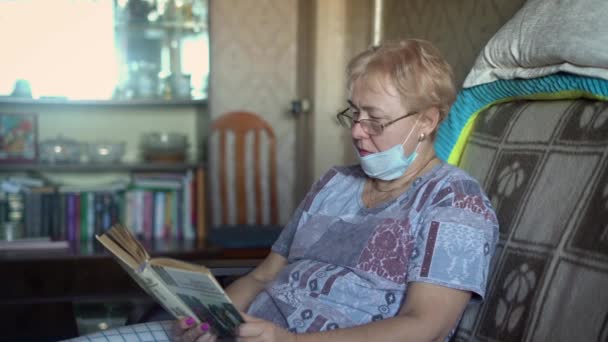 Oma met een masker die een boek leest. Ze is in zelfisolatie thuis tijdens de coronavirus pandemie. Ze verwacht dat een vrijwilliger haar eten brengt.. - Video