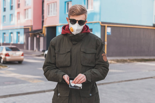 El hombre con un respirador de máscara protectora limpia su teléfono inteligente con una toallita antibacteriana húmeda, protegiéndose del virus. Medidas preventivas contra el coronavirus
. - Foto, Imagen