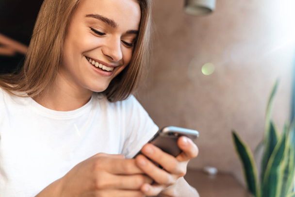 Photo of joyful young woman using cellphone and smiling while sitting in cozy cafe - Φωτογραφία, εικόνα
