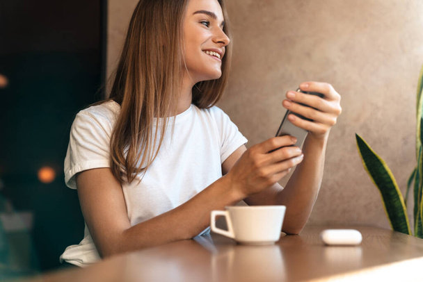 Photo of smiling young woman drinking coffee and using cellphone while sitting in cozy cafe - Foto, afbeelding