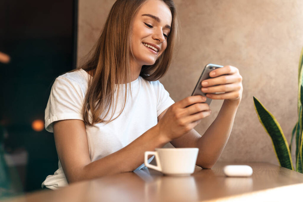 Photo of smiling young woman drinking coffee and using cellphone while sitting in cozy cafe - Foto, immagini