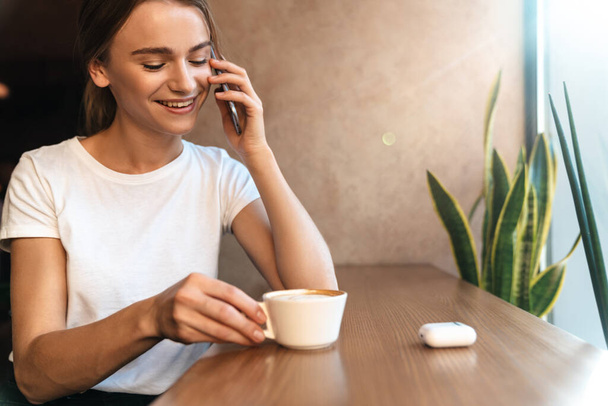 Foto einer lächelnden jungen Frau, die Kaffee trinkt und mit dem Handy telefoniert, während sie im gemütlichen Café sitzt - Foto, Bild