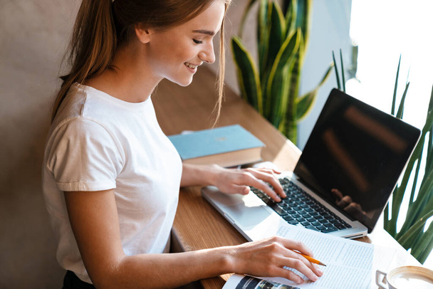 Gülümseyen güzel bir kadının bilgisayarı kullanırken ve rahat bir kafede otururken egzersiz kitabıyla çalışırken fotoğrafı. - Fotoğraf, Görsel