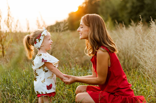 Kırmızı elbiseli çekici bir anne tarlada küçük kızının yanına çömelmiş. - Fotoğraf, Görsel