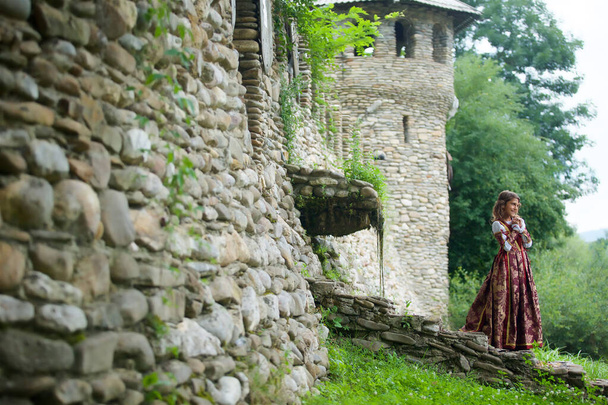 A little girl in a medieval red dress is located near the old castle with masonry. The child walks along the path near the building. - Фото, зображення