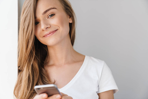 Photo of beautiful joyful woman in casual t-shirt using cellphone and smiling isolated over white wall - Foto, Bild