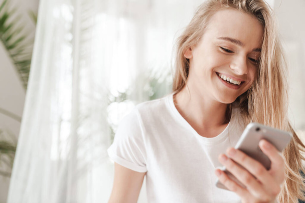 Imagem close-up de agradável jovem mulher com cabelo loiro digitando no smartphone e rindo no quarto ensolarado
 - Foto, Imagem