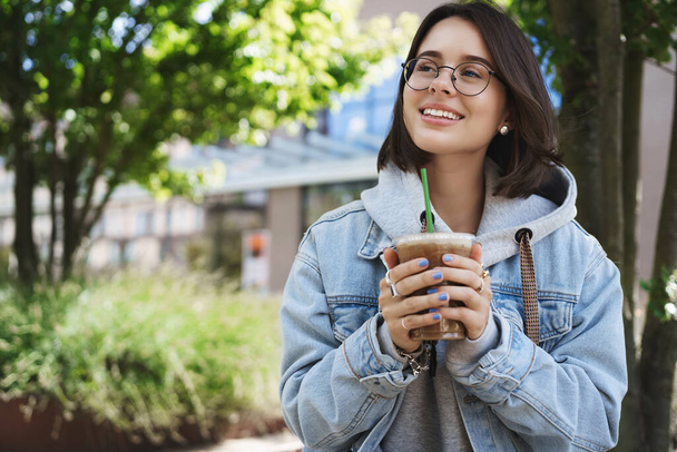 Portrait of young romantic and dreamy girl in glasses and denim jacket, looking up contemplating birds singing on bright perfect day, holding ice latte, drinking coffee and sit park bench smiling - Foto, Imagem