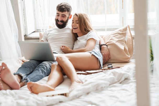 Image of romantic cheerful couple using laptop and hugging while lying on bed in bright room at home - Foto, afbeelding
