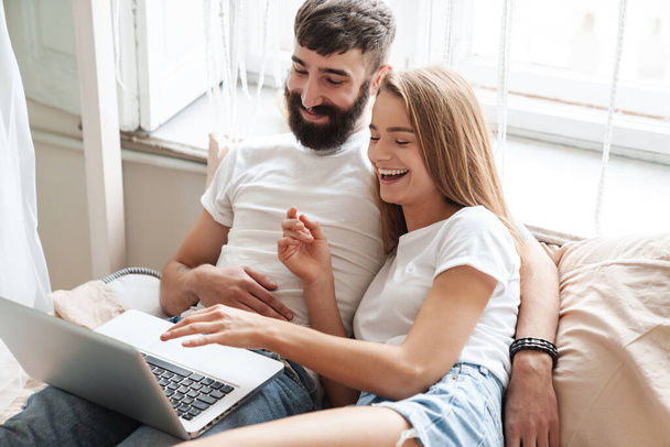 Image of nice excited couple using laptop and hugging while lying on bed in bright room at home - Foto, Bild