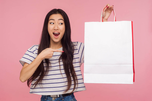 Portrait of surprised beautiful asian woman expressing amazement and pointing to shopping bag with white copy space, advertising area for text or image. indoor studio shot isolated on pink background - Фото, изображение