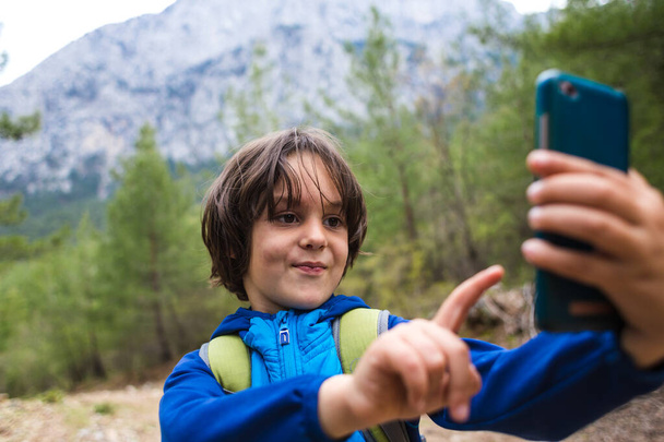 Ένα αγόρι με σακίδιο κάνει selfie σε ένα smartphone σε ένα φόντο από όμορφα βουνά. Παιδί σε εκδρομή κάμπινγκ. Το παιδί χρησιμοποιεί κινητό τηλέφωνο.. - Φωτογραφία, εικόνα