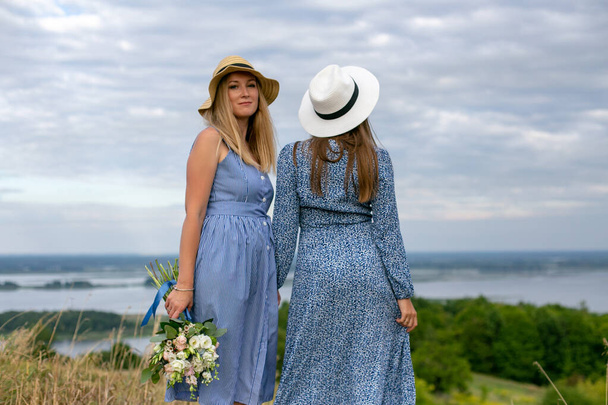 twee jonge langharige vrouwen in blauwe zomerjurken en hoeden staan op een heuvel boven de rivier te genieten van de prachtige natuur - Foto, afbeelding