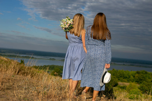 two beautiful long-haired young women in blue cotton dresses stand with their backs to the camera on the high bank of the river against the background of beautiful countryside - Photo, Image