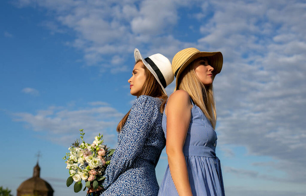 two young girls in blue summer dresses and hats stand with their backs to each other against a beautiful summer sky and the old church - Photo, Image