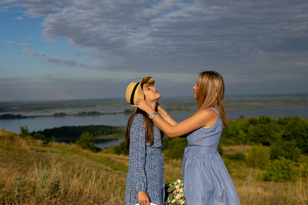 Дві дорослі дівчата-сестри стоять на тлі красивої дикої природи, обличчям один до одного, і один з них вирівнює капелюх іншого
 - Фото, зображення