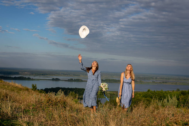 two happy young girls in blue summer dresses walk along the grassy high bank of the river and enjoy the beauty of wild untouched nature - Photo, Image