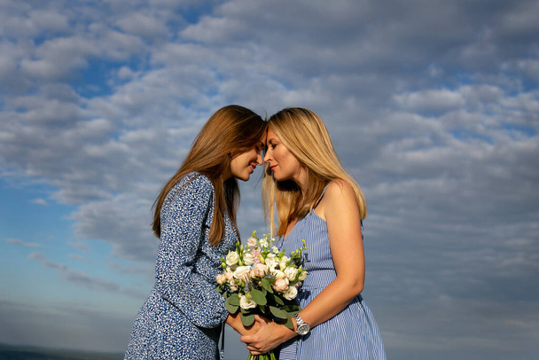 two adult girls sisters stand against a beautiful cloudy sky facing each other touching their foreheads - Photo, Image