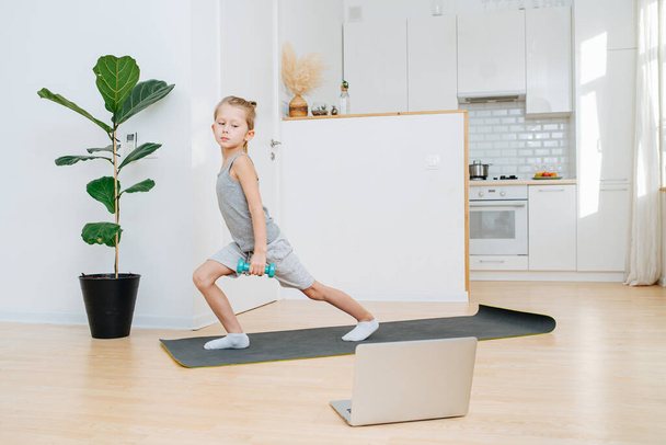 Boy schoolboy is doing sport online and performing exercise with dumbbells looking into laptop over the white kitchen - Foto, Imagen