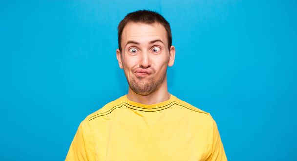 Αστείος κωμικός κάνει γκριμάτσα με σταυρωμένα μάτια. Νεαρός άνδρας σε κίτρινο t-shirt με τρελή έκφραση έχει τη διασκέδαση και μόνο, παίζει ανόητος απομονώνονται σε μπλε φόντο - Φωτογραφία, εικόνα