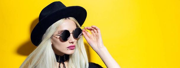 Portrét mladé blondýny nosí černý náhrdelník, klobouk a sluneční brýle na žlutém pozadí. - Fotografie, Obrázek