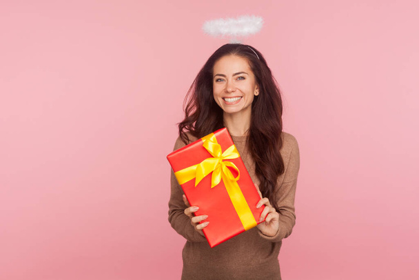 Portrait of happy beautiful angelic young woman with halo holding big present and smiling to camera, satisfied with gift, celebrating birthday, anniversary. studio shot isolated on pink background - 写真・画像