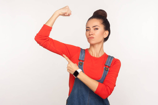 Look, I am independent and strong! Portrait of confident energetic stylish girl in denim overalls raising hand pointing at biceps, expressing power, female rights. indoor studio shot, white background - Фото, изображение