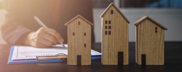Концепція підписання контракту на нерухомість, невелика дерев'яна модель будинку на офісному столі з рукою жінки-покупця знак на папері договору оренди для оренди вище зазначеного місця проживання
 - Фото, зображення