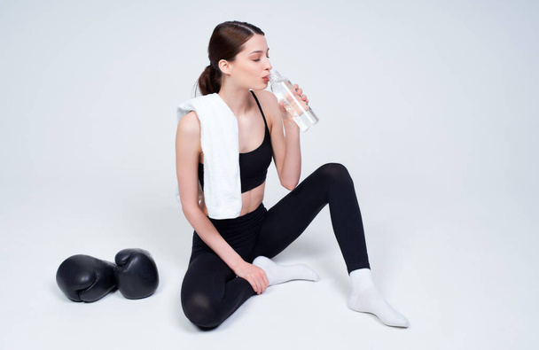 Charming romantic tender young girl, in a black sports top and leggings, enjoys drinking water after a workout, boxing gloves lie nearby. Sports girl resting after class on a white background. - Fotó, kép