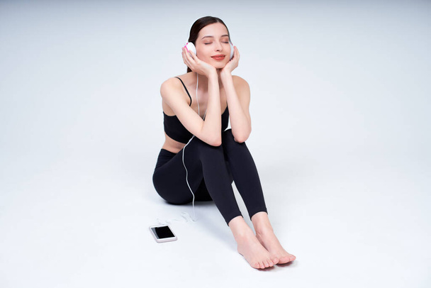 Charming cute young fresh Caucasian girl in a black sports suit for sports, sitting on a white background, listening to music on headphones, resting after a workout, dreamily closed her eyes. - Photo, image