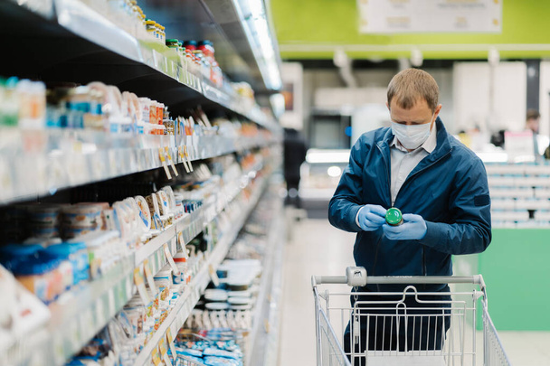 Horizontale shot van volwassen man draagt beschermende masker, leest etiket van het product, maakt winkelen tijdens coronavirus uitbraak, koopt nessecary voedsel in de lokale winkel. Mensen, virus, ziekte, aankoopconcept - Foto, afbeelding
