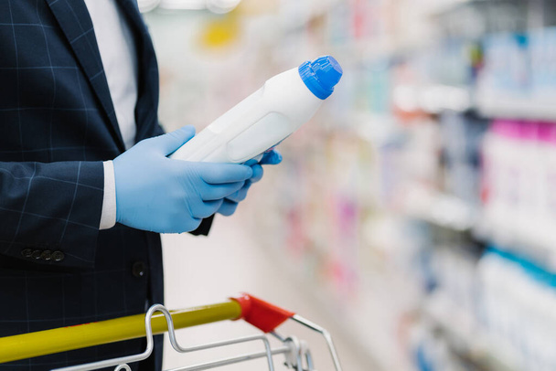 Man chooses detergent in household goods store, holds bottle with liquid powder, wears medical gloves to protect from coronavirus, reads product information. Making shopping during quarantine - Photo, Image