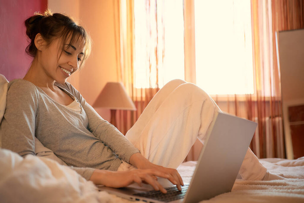 mujer sonriente satisfecho en la cama en casa charlando con amigos en las redes sociales en el ordenador portátil, el uso de Internet velocidad inalámbrica. Mujer comunicándose a través de chat de vídeo en línea, escribiendo en el teclado del ordenador portátil
. - Foto, imagen