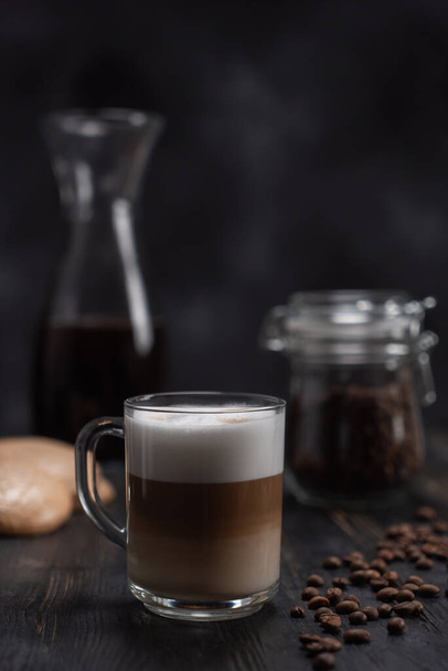 Transparent Cup of coffee cappuccino or latte is on the black table next to cookies and a jar of coffee beans. A Cup of coffee cappuccino or latte with cinnamon and coffee beans on a black wooden - Foto, Bild