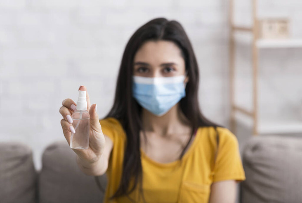 COVID-19 prevention. Girl in protective mask holding bottle of sanitizer at home, selective focus - Φωτογραφία, εικόνα