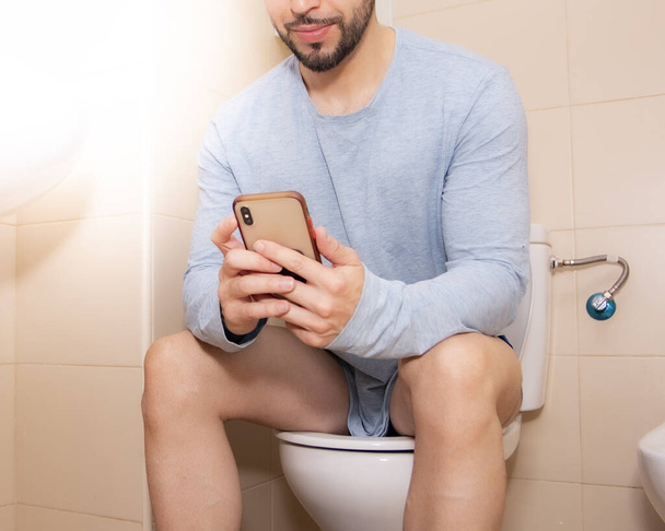 ενήλικας άνδρας που χρησιμοποιεί smartphone ενώ κάθεται σε λεκάνη τουαλέτας στο σπίτι, έννοια της τεχνολογίας και του τρόπου ζωής β - Φωτογραφία, εικόνα