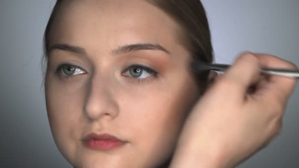 Макияж художник делает профессиональный макияж для молодой женщины в студии красоты. Художник наносит румяна кистью на веки - Кадры, видео