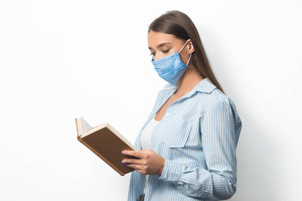 Κορίτσι ανάγνωση βιβλίο φορώντας ιατρική μάσκα πάνω από το λευκό φόντο στούντιο - Φωτογραφία, εικόνα