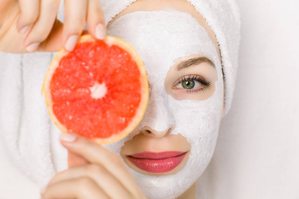 Close up of beautiful young smiling woman with towel on head, facial skin mask, holding grapefruit slice in front of her eye, lying on white background. Top view, focus on face - Fotografie, Obrázek